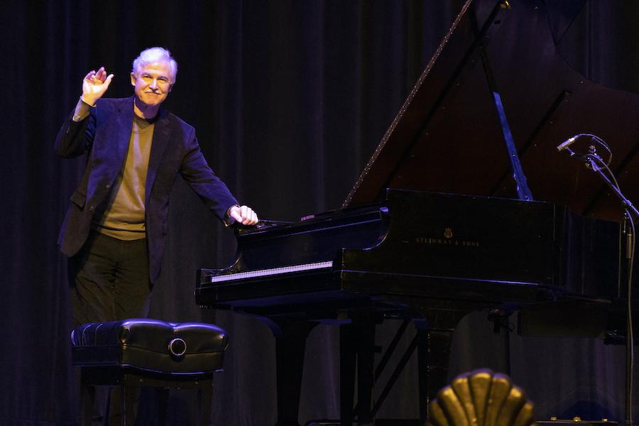 A grey-haired man in his 70, standing next to a black grad piano, waves to an audience following his performance.