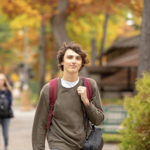 A smiling student wearing a green shirt strides down a sidewalk on Interlochen's campus.